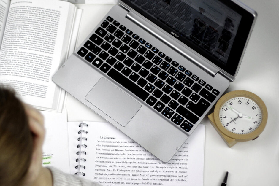 Eine Studentin sitzt an einem Schreibtisch, auf dem ein Laptop, ein Wecker, auf aufgeschlagenes Buch und ein Vorlesungsskript liegen.