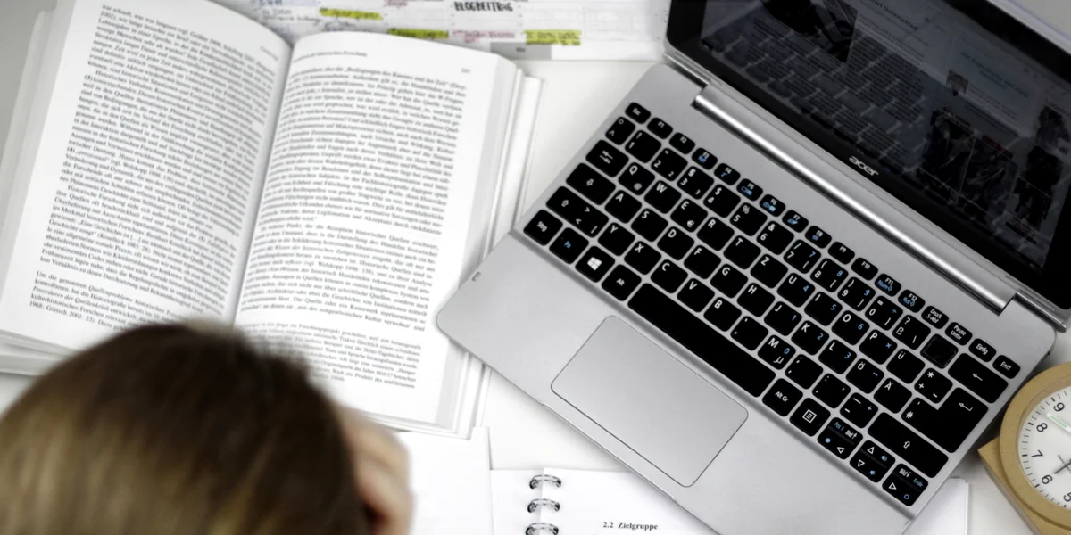 Eine Studentin sitzt an einem Schreibtisch, auf dem ein Laptop, ein Wecker, auf aufgeschlagenes Buch und ein Vorlesungsskript liegen.