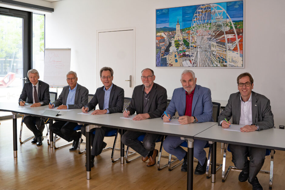 Sechs Männer in formeller Kleidung stitzen an einem langen Tisch in einem Seminarraum. Vor ihnen liegt jeweils ein Stapel Papiere, alle halten einen Stift in der rechten Hand und blicken lächelnd in die Kamera. (öffnet Vergrößerung des Bildes)