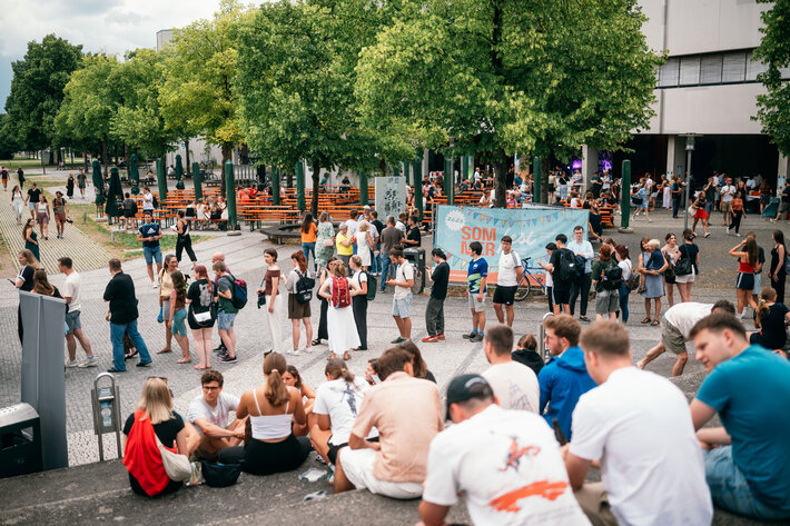Besucherinnen und Besucher des Uni-Sommerfests sitzen im Freien vor dem PT-Gebäude. (öffnet Vergrößerung des Bildes)