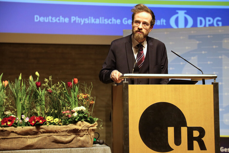 Ein Mann im Anzug, mit Brille und Bart steht hinter einem mit UR-Logo geschmückten Rednerpult.