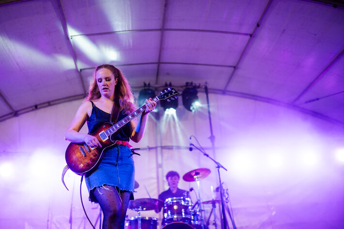 Eine junge Frau mit langen blonden Haaren, schwarzem Oberteil und Jeansrock spielt Gitarre. Im Hintergrund sieht man das Schlagzeug mit seiner Spielerin. Die Bühnenbeleuchtung ist violett. (öffnet Vergrößerung des Bildes)