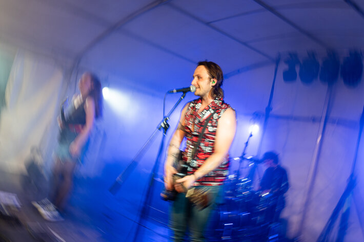 Ein junger Mann in bunter Weste und Jeans steht am Mikrofon, singt und spielt Gitarre. Die Bandkolleginnen im Hintergrund sind verschwommen. Die Bühnenbeleuchtung ist blau. (öffnet Vergrößerung des Bildes)