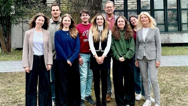 9 Personen stehen an einem grauen Tag vor einem Baum vor einem Gebäude auf dem Campus. Es handelt sich um das Gruppenfoto des Lehrstuhlteams.
