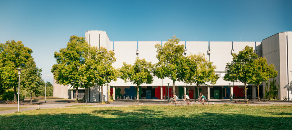 Drei junge Personen auf Fahrrädern vor einem Gebäude mit großen, strahlend hellen Betonflächen in der späten Nachmittagssonne. Der Himmel ist Tiefblau, das Gebäude ist umgeben von rünen Bäumen und einer grünen, mit Wiesenblumen gesprenkelten Wiese.