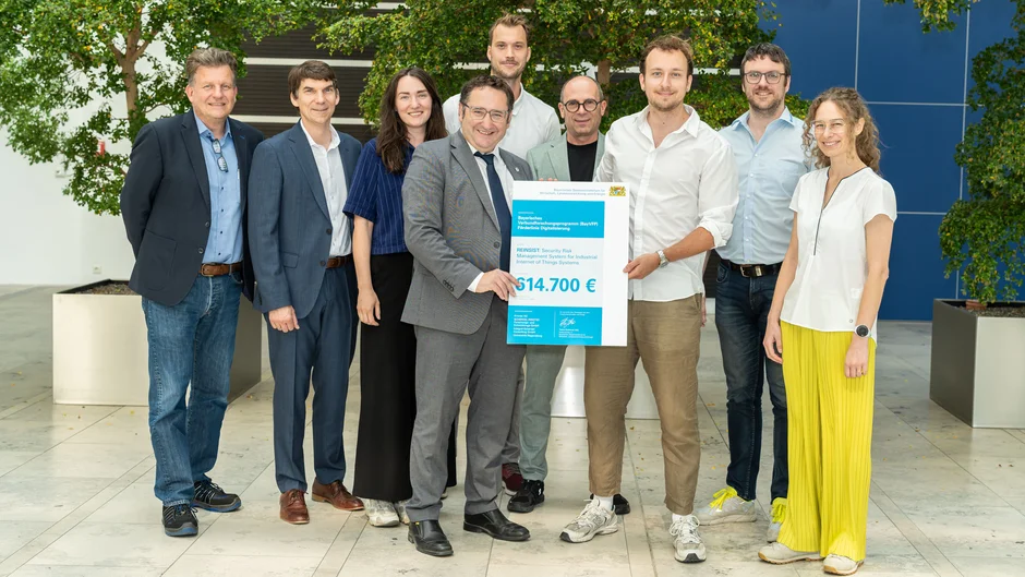 Neun Personen präsentieren in einer modernen, hellen Halle mit großen Bäumen eine Förderurkunde für das Forschungsprojekt „REINSIST“. Im Mittelpunkt wird ein Förderscheck des Bayerischen Verbundforschungsprogramms über 614.700 Euro gehalten. (öffnet Vergrößerung des Bildes)