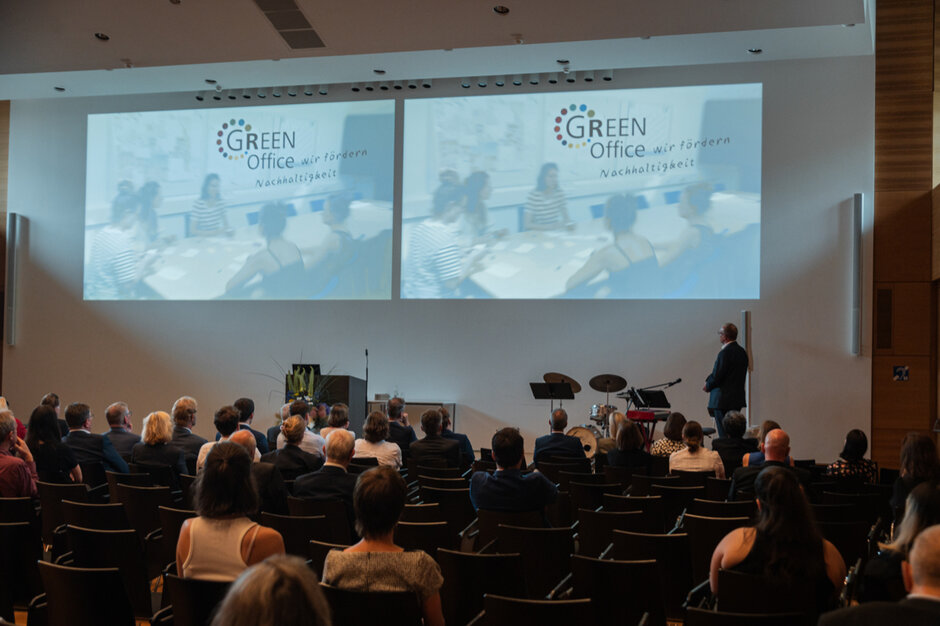 Blick in einen Hörsaal. Mehrere Stuhlreihen stehen hintereinander, auf vielen Stühlen sitzen Personen mit Blick zur Bühne. Auf der Bühne steht ein Mann und hat sich Richtung Wand gewendet, auf der eine gedoppelte Projektion mit dem Titel "Green Office" zu sehen ist.