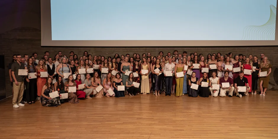 Großes Gruppenfoto auf einer Bühne mit Holzfußboden. Mehr als 100 Personen sind in mehreren Reihen angeordnet, einige stehen, andere sitzen oder knien in der ersten Reihe. Viele halten Urkunden und einzelne Blumen in den Händen. Die Teilnehmenden tragen festliche Kleidung. Über der Gruppe befindet sich eine große Projektionsfläche. (öffnet Vergrößerung des Bildes)