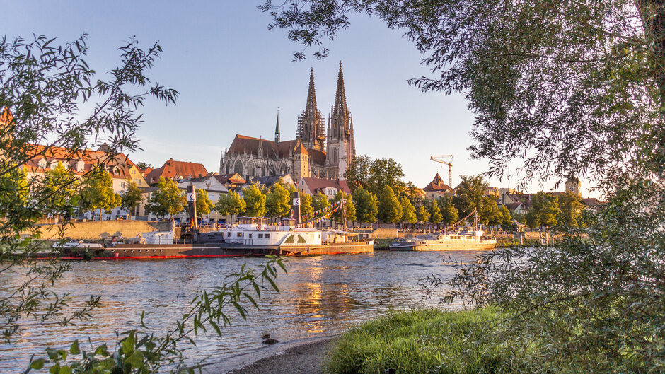 Blick auf die Donau, das südliche Ufer, Ausflugsschiffe und die Regensburger Altstadt. Im Hintergrund die Domspitzen.