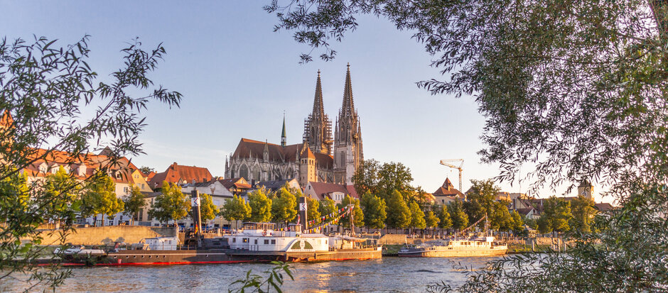 Blick auf die Donau, das südliche Ufer, Ausflugsschiffe und die Regensburger Altstadt. Im Hintergrund die Domspitzen.