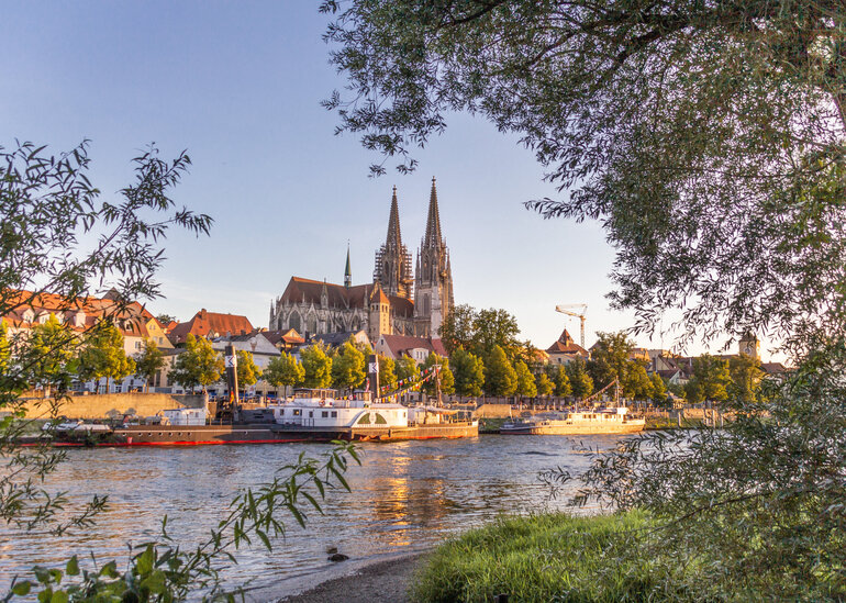Blick auf die Donau, das südliche Ufer, Ausflugsschiffe und die Regensburger Altstadt. Im Hintergrund die Domspitzen.