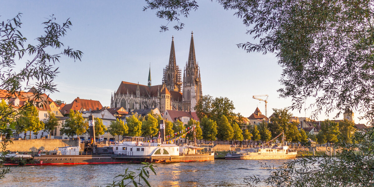 Blick auf die Donau, das südliche Ufer, Ausflugsschiffe und die Regensburger Altstadt. Im Hintergrund die Domspitzen.