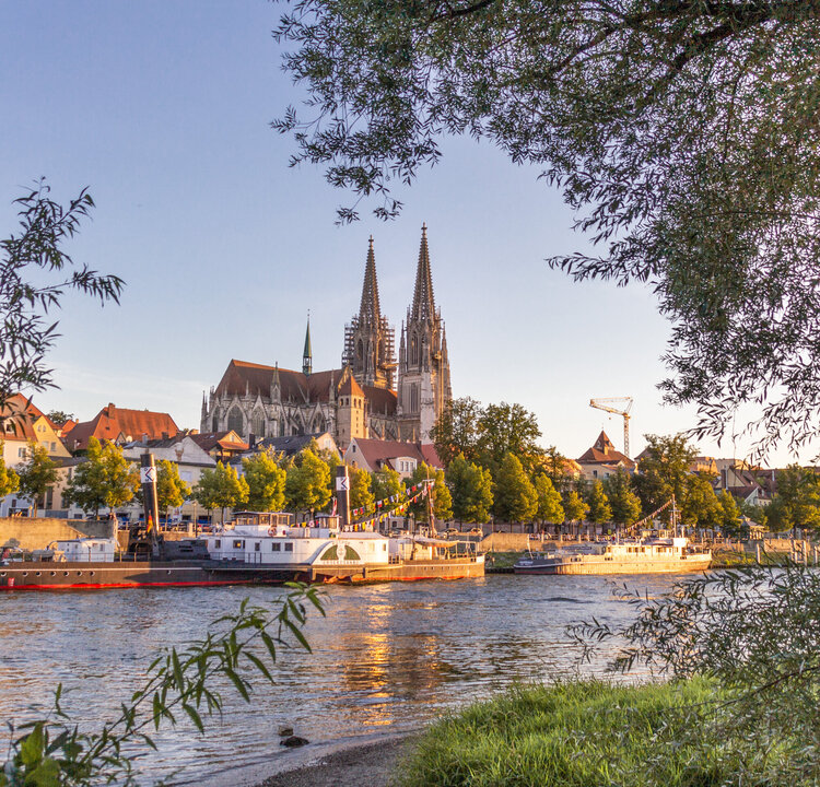 Blick auf die Donau, das südliche Ufer, Ausflugsschiffe und die Regensburger Altstadt. Im Hintergrund die Domspitzen.