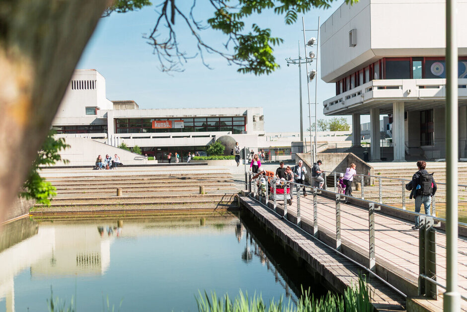 Der See der Uni Regensburg mit einer Brücke, auf der einige Menschen zu sehen sind. Im Hintergrund sieht man das Audimax.