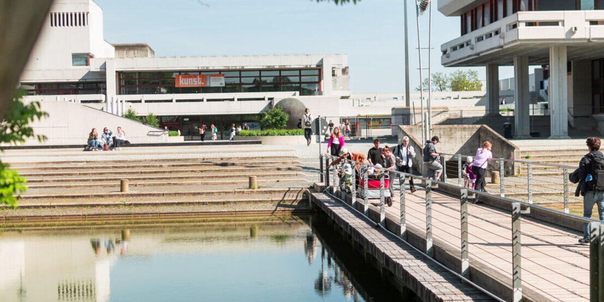 Der See der Uni Regensburg mit einer Brücke, auf der einige Menschen zu sehen sind. Im Hintergrund sieht man das Audimax.
