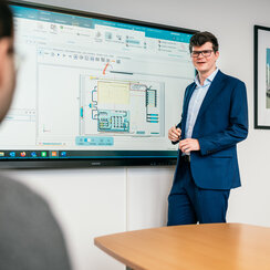 Ein Professor steht an einer elektronischen Tafel, auf der ein Schaubild zu sehen ist. Er spricht mit einer vor ihm sitzenden Person.
