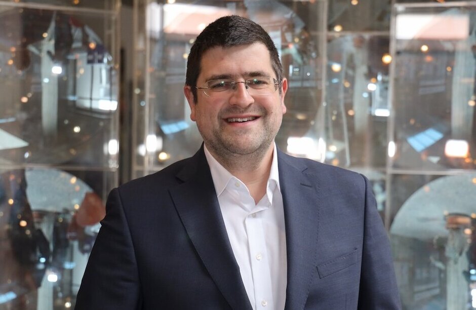 Porträtfoto: Mann mit Brille und kurzen, dunklen Haaren vor neutralem Hintergrund