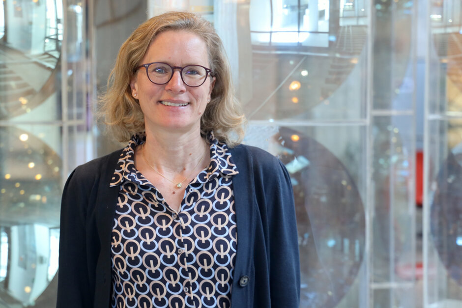 Eine Frau mit blonden, gewellten Haaren und Brille blickt lächelnd Richtung Kamera.