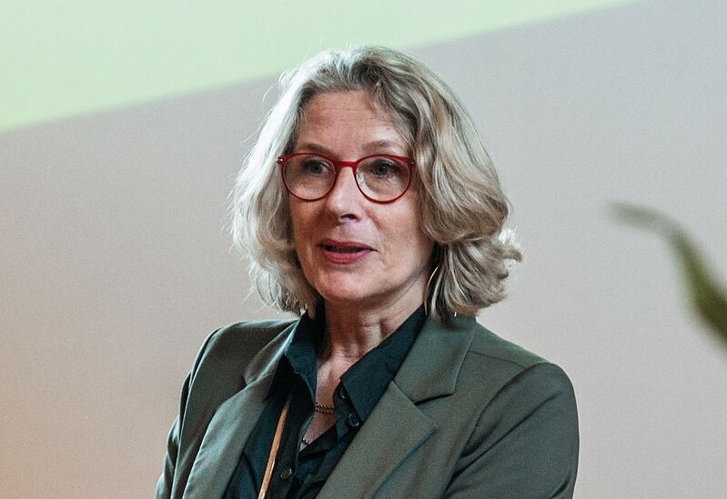 Eine Frau mit grauen Haaren und Brille mit rotem Brillengestell spricht.