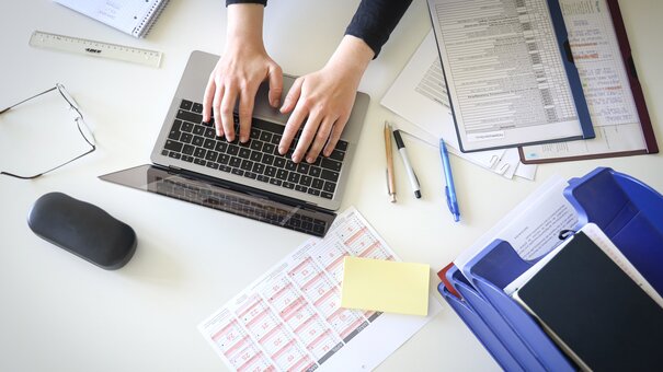 Man sieht Hände, die an einem Notebook arbeiten. Es liegen Papiere und ein Handy verteilt auf dem einem weißen Tisch.