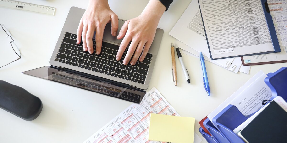 Man sieht Hände, die an einem Notebook arbeiten. Es liegen Papiere und ein Handy verteilt auf dem einem weißen Tisch.
