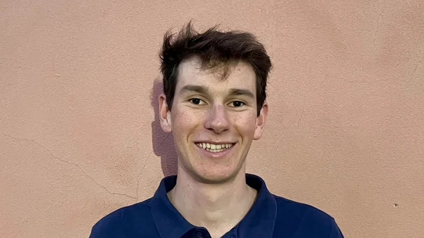 A portrait photograph of a smiling young man with short brown hair, wearing a dark blue shirt and standing in front of an ochre-coloured wall.
