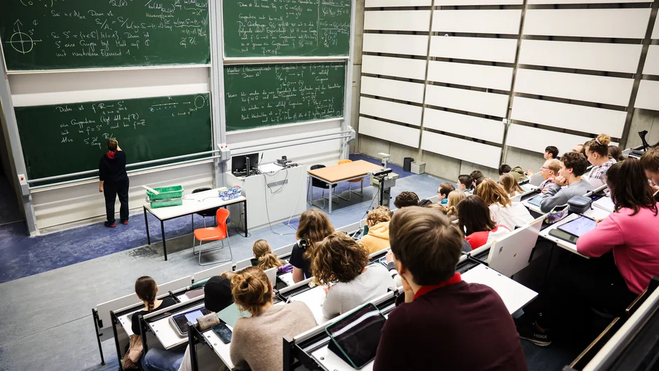 Studierende in Hörsaal, Frau schreibt Formeln an Tafel. (öffnet Vergrößerung des Bildes)