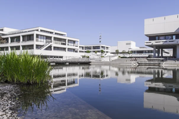 Universitätscampus mit Gebäuden; in der Mitte ein See, in dem sich die Fassaden und der blaue Himmel spiegeln.