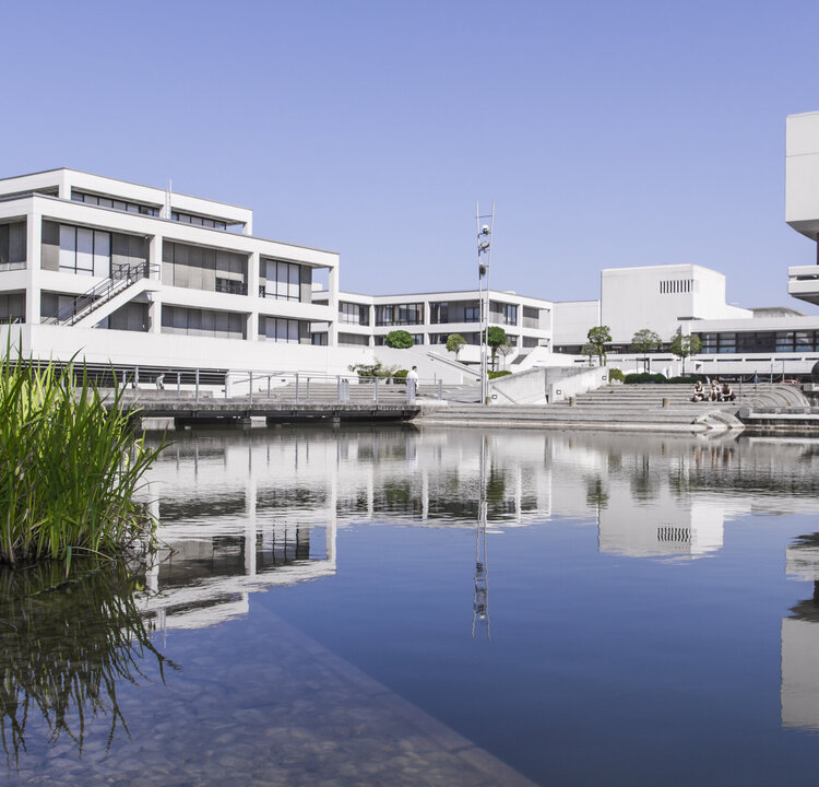Universitätscampus mit Gebäuden; in der Mitte ein See, in dem sich die Fassaden und der blaue Himmel spiegeln.