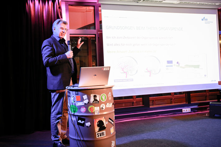 Ein Vortragender steht auf einer Bühne neben einer großen Projektionsfläche, hält ein Mikrofon und gestikuliert mit der freien Hand. Neben ihm steht ein Laptop auf einem Fass, das mit Aufklebern beklebt ist. Auf der Präsentationsfolie ist die Überschrift „GRUNDSORGEN BEIM THEMA ORGANSPENDE” sowie Fragen zur Organspende in deutscher Sprache zu sehen. Die Szene ist in violett‑rotes Bühnenlicht getaucht, im Hintergrund sind Vorhänge und Fenster sichtbar. (öffnet Vergrößerung des Bildes)