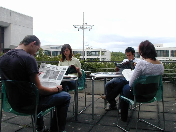 Vier Personen sitzen lesend auf der Leseterrasse, im Hintergrund Gebäude (öffnet Vergrößerung des Bildes)
