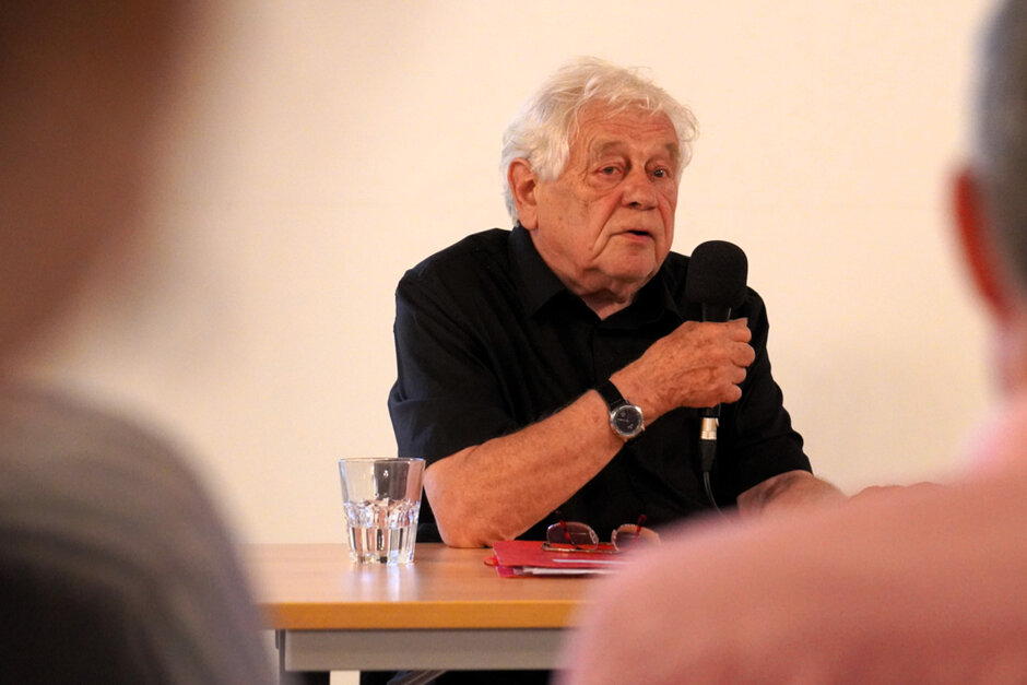 Ein älterer Mann in einem kurzärmligen schwarzen Hemd hält ein Mikrofon in der Hand, in das er spricht. (öffnet Vergrößerung des Bildes)