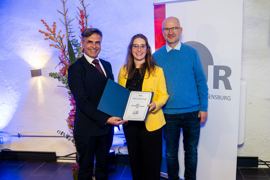Zwei Männer und eine Frau stehen vor einem Rollup, das das Logo der Universität Regensburg und einen Farbbalken abbildet. Im Hintergrund sieht man ein Blumengesteck. Die Frau im gelben Blazer hält eine Urkunde in die Kamera.