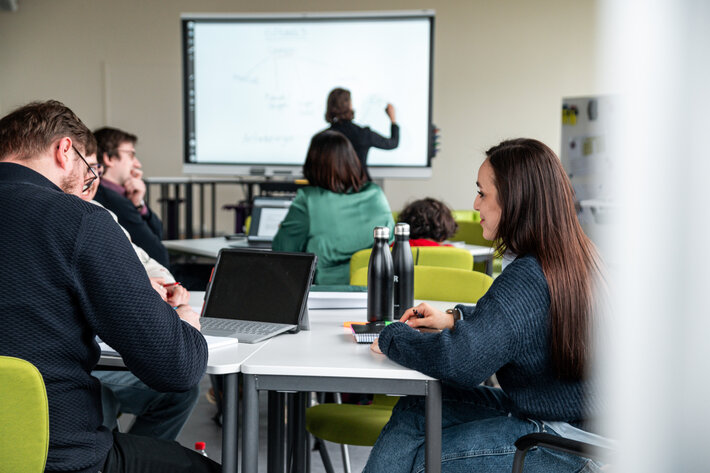 Studierende sitzen in einem Seminarraum, im Hintergrund schreibt eine Dozentin an ein interaktives Whiteboard. (öffnet Vergrößerung des Bildes)