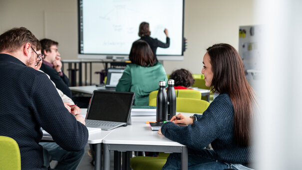 Studierende sitzen in einem Seminarraum, im Hintergrund schreibt eine Dozentin an ein interaktives Whiteboard.