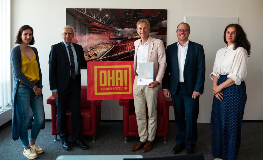 Fünf Personen stehen zusammen in einem Büro. Zwie Männer halten ein Schild mit dem Schriftzug „OHA OSTBAYERN HANDELT!“. Einer von den beiden hält außerdem eine Urkundenmappe in der Hand. (öffnet Vergrößerung des Bildes)