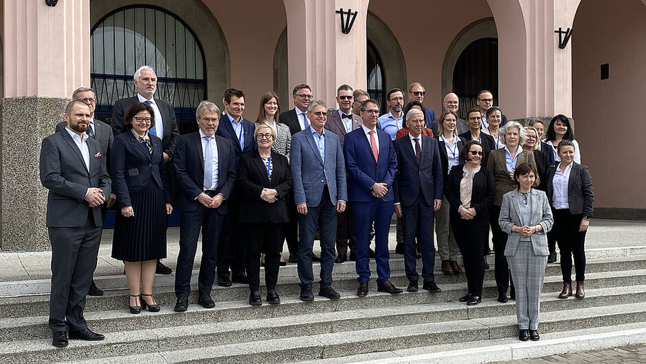 Gruppenbild mit rund 20 Personen in formeller Kleidung vor dme Eingangsportal zu einem öffentlichen Gebäude.