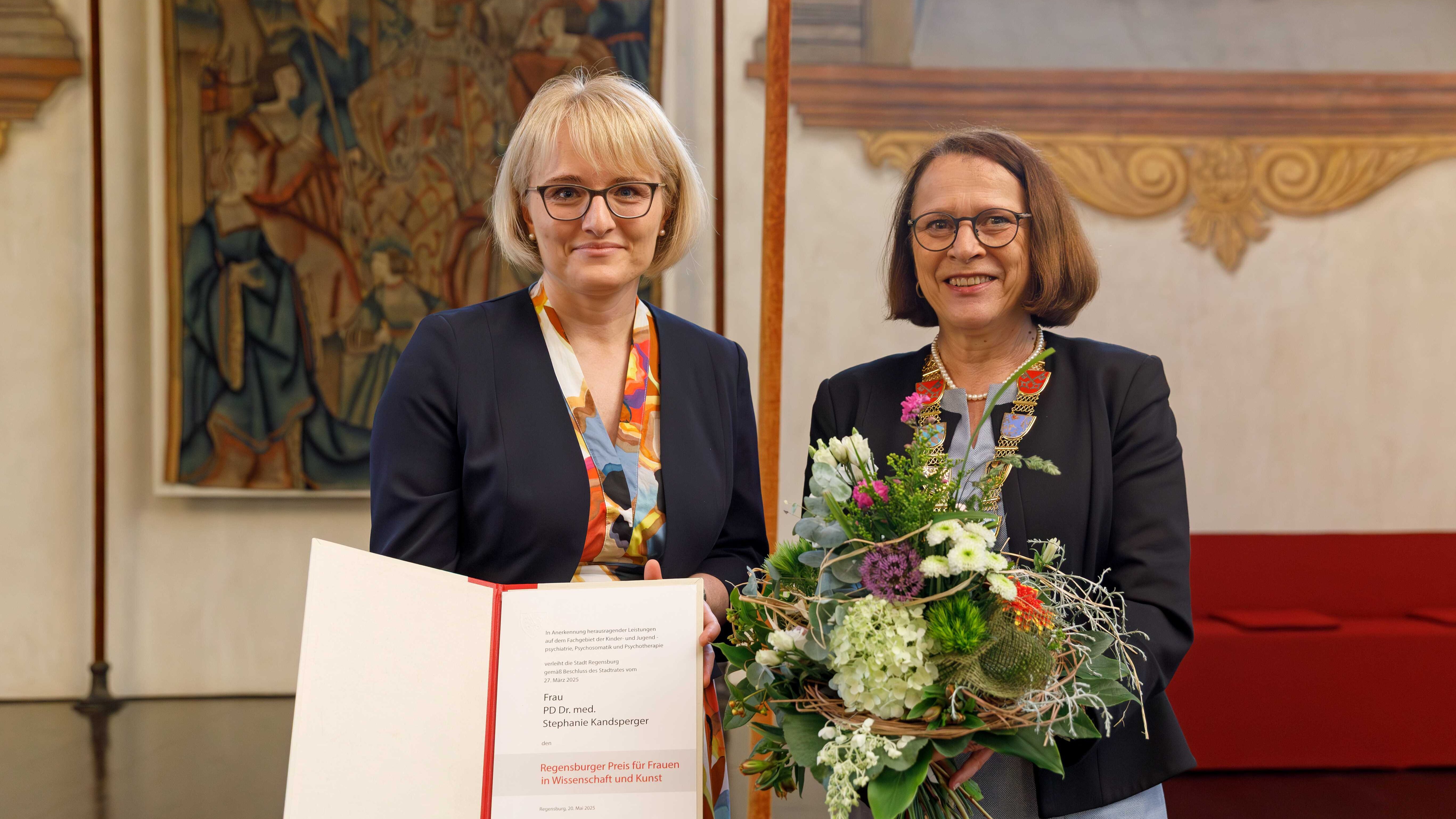 Auf dem Bild stehen zwei Frauen und schauen in die Kamera. Die eine Frau hat blonde Haare, trägt eine Brille und trägt ein buntes Kleid mit einem schwarzen Blazer. In der Hand hält sie eine Urkunde. Die andere Frau hat braune Haare, trägt eine Brille, hellblaues Kleid und einen schwarzen Blazer. In der Hand hält sie einen bunten Blumenstrauß. (öffnet Vergrößerung des Bildes)