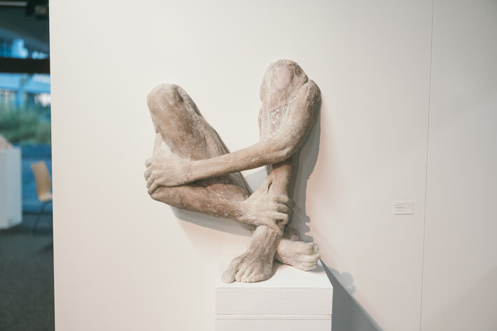 Das Bild zeigt eine Plastik: zwei menschliche Beine vom Knie abwärts werden von zwei menschlichen Armen vom Ellenbogen abwärts umschlungen. Die vier Gliedmaßen scheinen aus der Wand zu kommen. (öffnet Vergrößerung des Bildes)