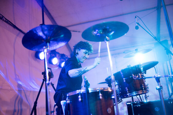 Eine junge Frau mit kurzen Haaren und tätowiertem Unterarm in schwarzem kurzärmeligem Hemd sitzt am Schlagzeug und spielt darauf. Die Bühnenbeleuchtung ist blau-rot. (öffnet Vergrößerung des Bildes)