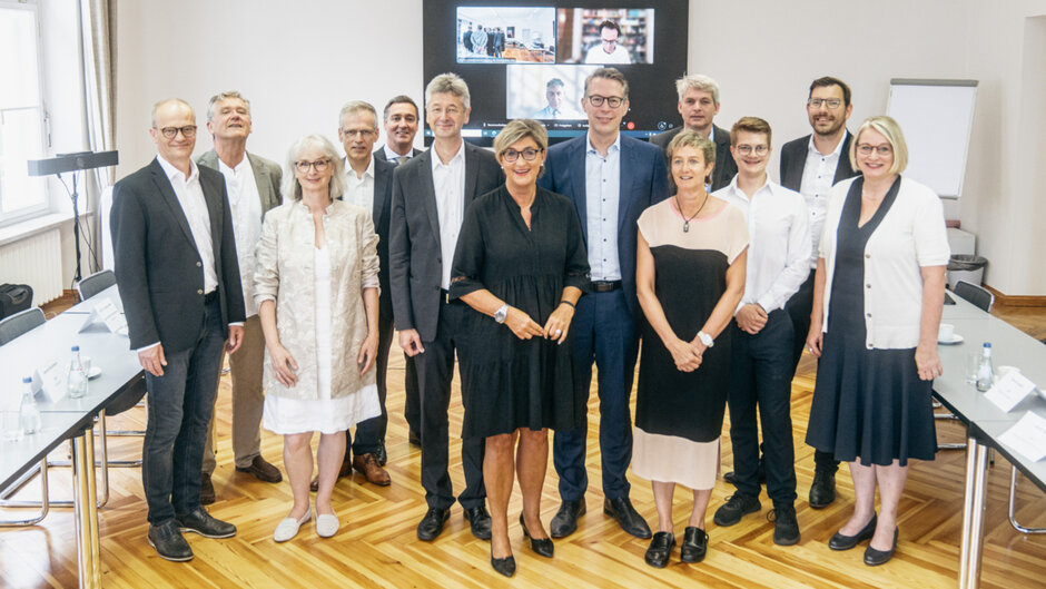 Gruppenbild mit mehreren Personen in formeller Kleidung, die sich für ein Foto in der Mitte eines Konferenzraumes aufgestellt haben. (öffnet Vergrößerung des Bildes)