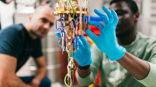 Ein physikalischer Versuchsaufbau mit Kabeln, Kupferelementen etc. Ein Mann mit blauen Einmalhandschuhen nimmt letzte Justierungen vor. Im Hintergrund blickt ein weiterer Mann interessiert auf den Versuchsaufbau.