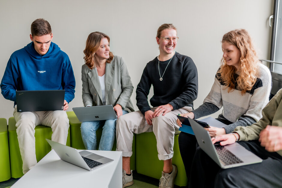 Eine Gruppe junger Leute sitzt auf hellgrünen Hockern und unterhält sich. Manche von ihnen haben Laptops auf den Knien.