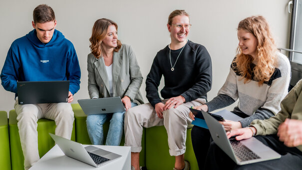 Eine Gruppe junger Leute sitzt auf hellgrünen Hockern und unterhält sich. Manche von ihnen haben Laptops auf den Knien.