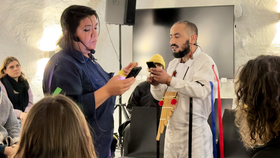 Eine Frau in einem blauen Overall mit einem Headset liest etwas von einem Smarphone ab. Rechts neben ihr steht ein Mann in einem weißen Overall mit einer Panflöte um den Hals und bunten Bändern an seinem linken Ärmel. Er scheint etwas von einem Smartphone vorzulesen.