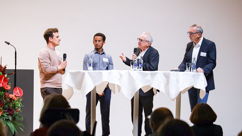 Vier Männer an Stehtischen, einer hält Mikrofon. (öffnet Vergrößerung des Bildes)