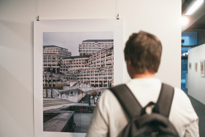 Ein Veranstaltungsbesucher betrachtet ein ausgestelltes Kunstwerk. Auf dem Kunstwerk sind Fotos von Betonbauten komponiert. (öffnet Vergrößerung des Bildes)