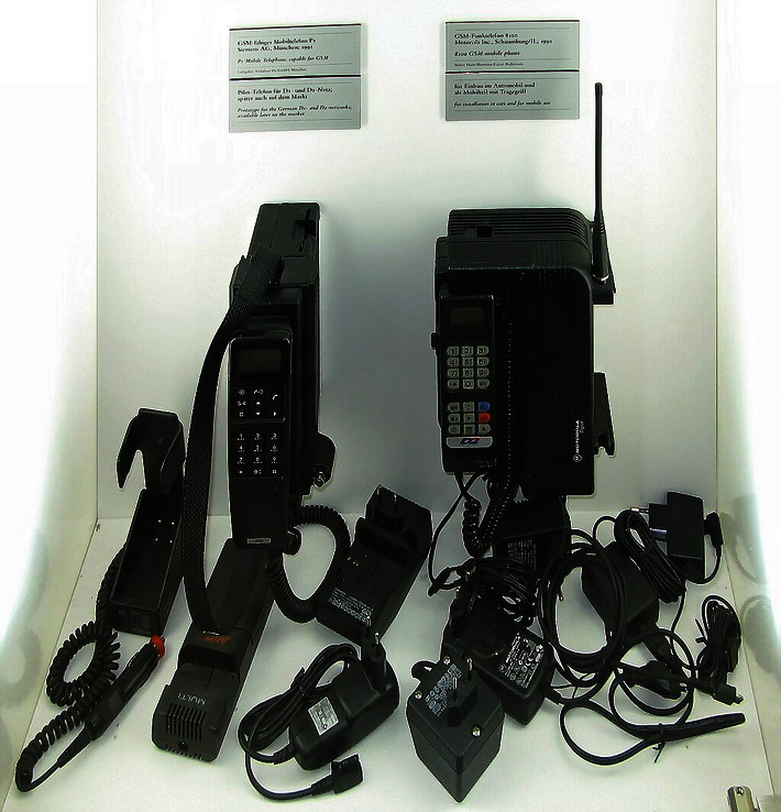 SM-fähige Mobiltelefone ([links:] P1 der Siemens AG, M¸nchen, 1991; [rechts:] Motorola 8100, 1991) mit Zusatzakku, Auto- und Stromnetzladeeinheiten, Wandhalterungen (öffnet Vergrößerung des Bildes)