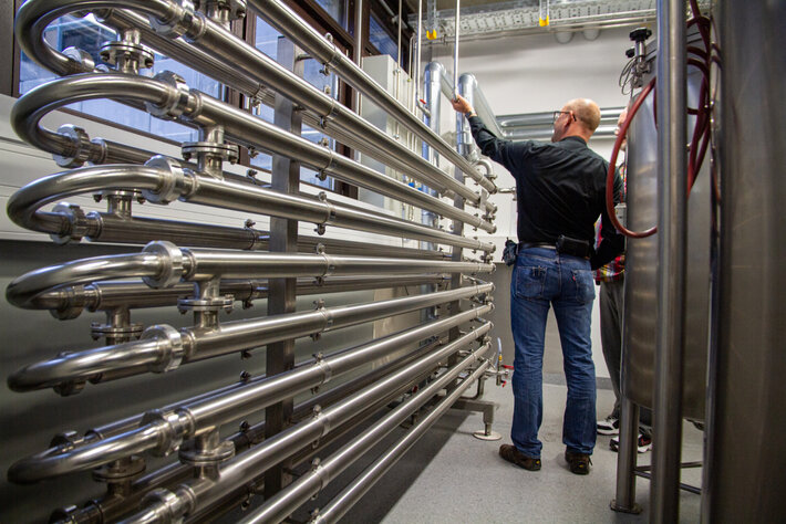 Personen betrachten eine große technische Anlage mit mehreren parallel verlaufenden Edelstahlrohren in einem Laborraum. (öffnet Vergrößerung des Bildes)