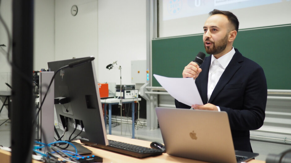 Ein Mann in weißem Hemd und schwarzem Anzug steht am Rednerpult in einem Hörsaal und spricht in ein Mikrofon.